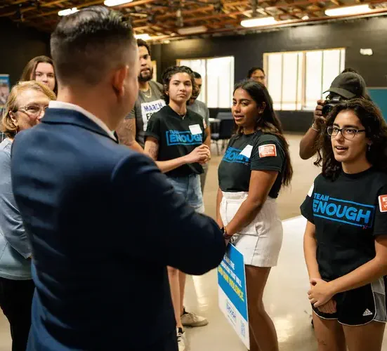 A group of people wearing t-shirts that read "Team Enough" looking at aa man in a navy blue suit.